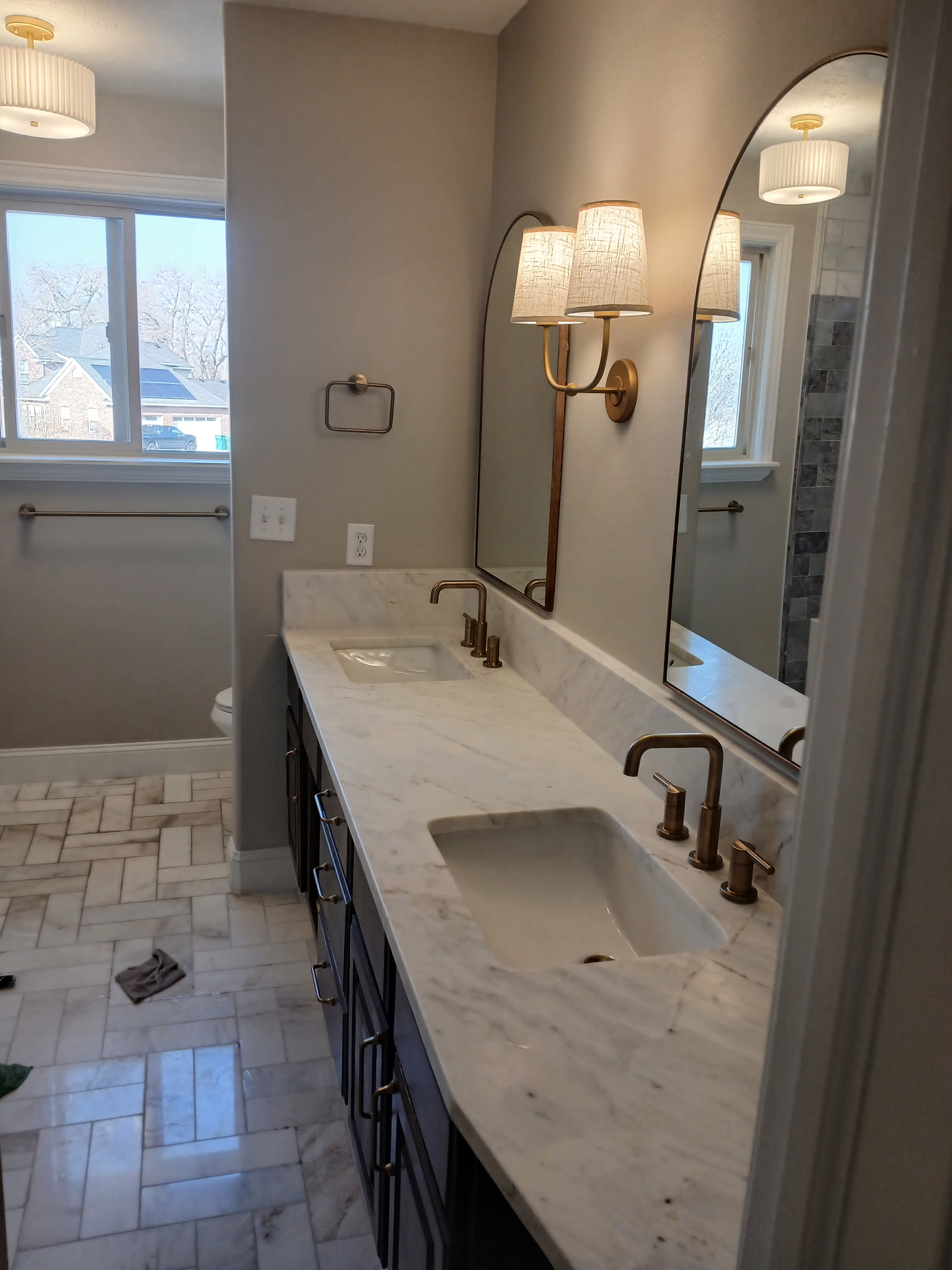 Luxury master bathroom with double marble vanity, brass sconces, arched mirrors, and herringbone marble floor tile